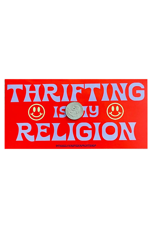 Red bumper sticker on white background. The sticker says Thrifting is my Religion. On either side of the text are two yellow smiley faces. On top of the sticker is a quarter to show the sticker size.