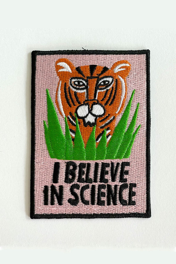 A vertical rectangular embroidered patch with a black border and black text that reads "I believe in science." The interior of the patch is light pink with an illustration of an orange tiger's head hiding in tall blades of green grass. 