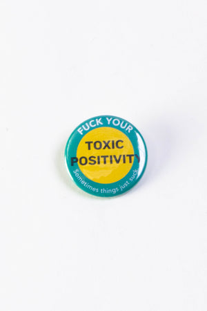 Round pinback button on a white background. The button has a blue green border around it with a large. yellow dot in the middle. The button says "Fuck Your Toxic Positivity. Sometimes things just suck." The text is white, with the exception of Toxic Positivity which is in a black font.