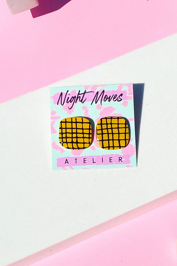 A pair of rounded square mustard yellow earrings with a black asymmetrical grid pattern. The earrings are attached to a teal and pink backing that has the brand name "Night Moves Atelier."