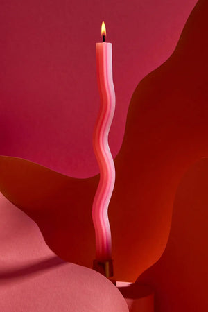 A lit thin and tall orange and pink squiggly shaped votive candle in front of a red background of various shapes and shades of red. Metal stand not included.