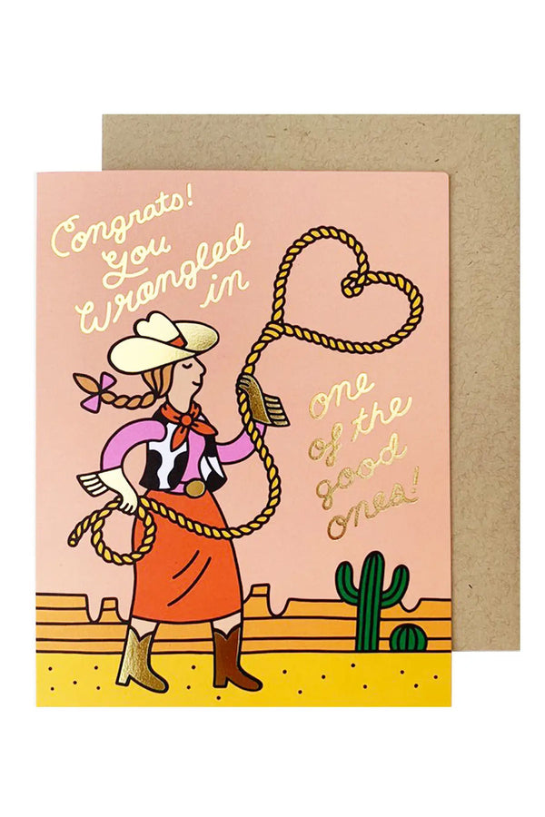 A greeting card with an illustration of a cowgirl swinging a lasso. She's wearing a tan cowboy hat with her hair in a braid, a pink shirt, red bandana around her neck, and a cow-print vest with a red skirt and boots. She is standing in the foreground of a desert landscape. The gold cursive text reads "Congrats! You wrangled in one of the good ones!"